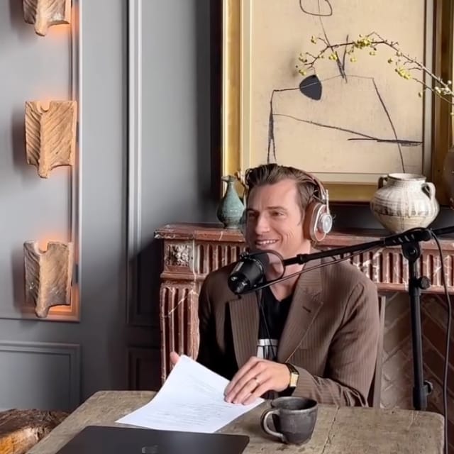 A podcast host in a brown blazer and headphones speaks into a microphone while holding notes. The setup includes a laptop and coffee mug on the table, with an ornate fireplace, decorative vases, and abstract wall art in the background.